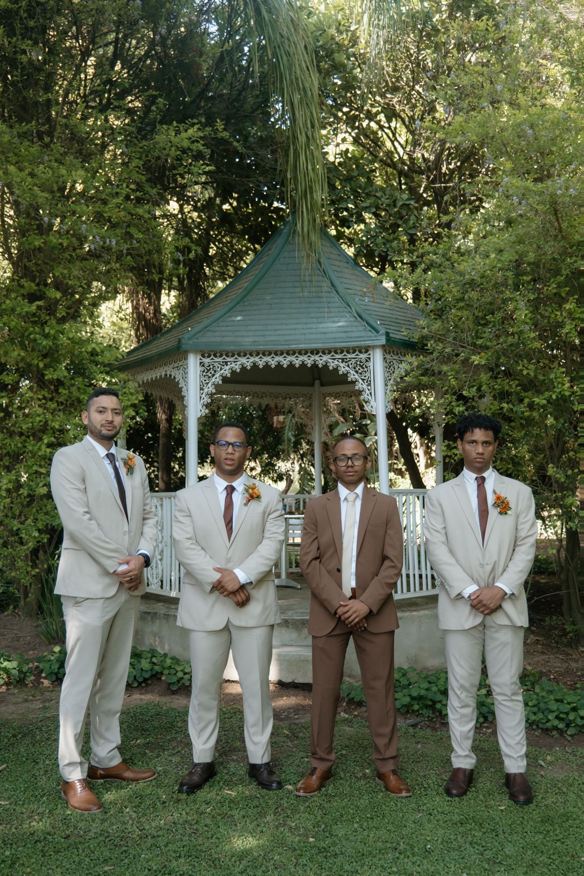 Four groomsmen in beige and brown suits pose in front of a white gazebo surrounded by greenery.