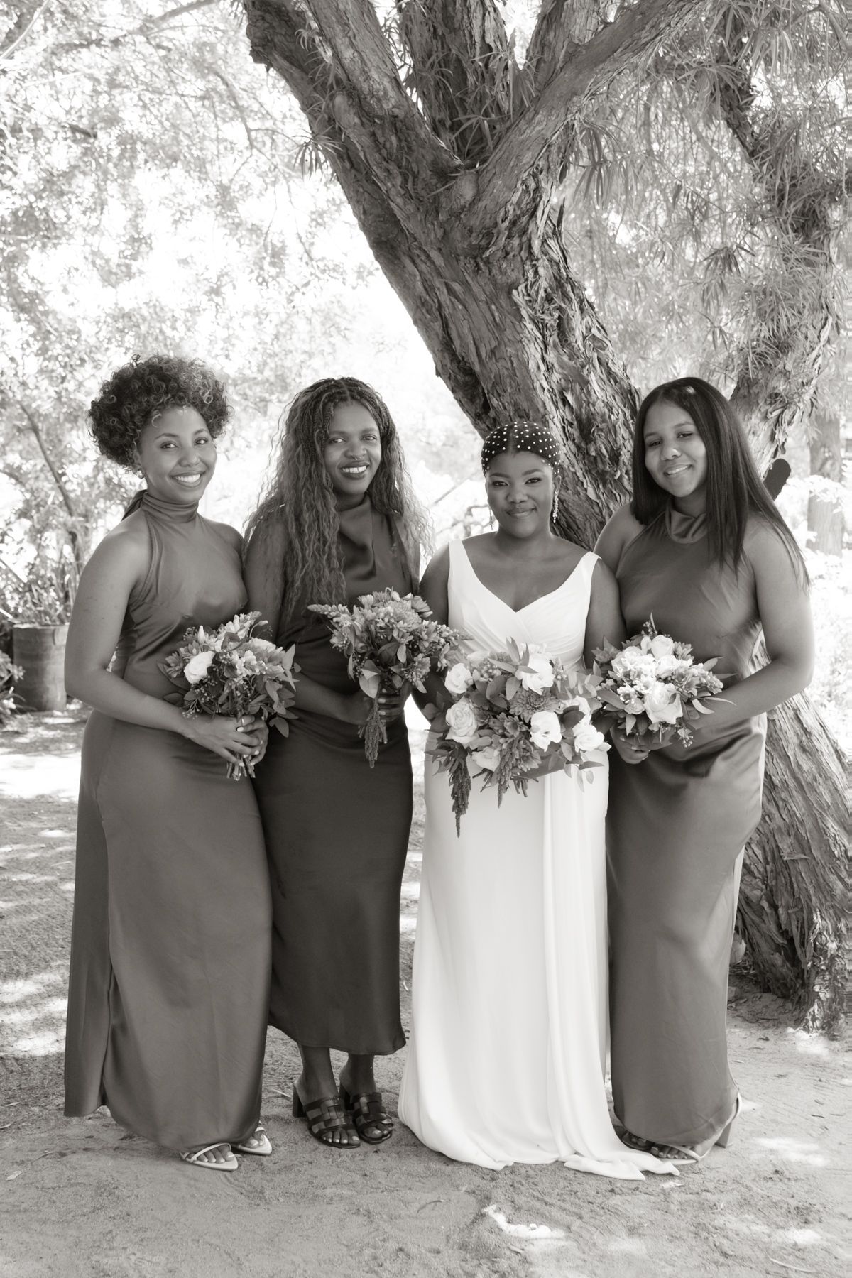 Black and white photo of bride in white gown with three bridesmaids in elegant dresses holding floral bouquets outdoors.