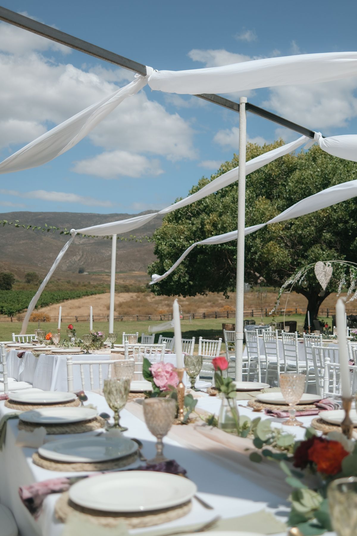 Outdoor wedding reception tables with white draping canopy, floral centerpieces, and scenic mountain landscape.