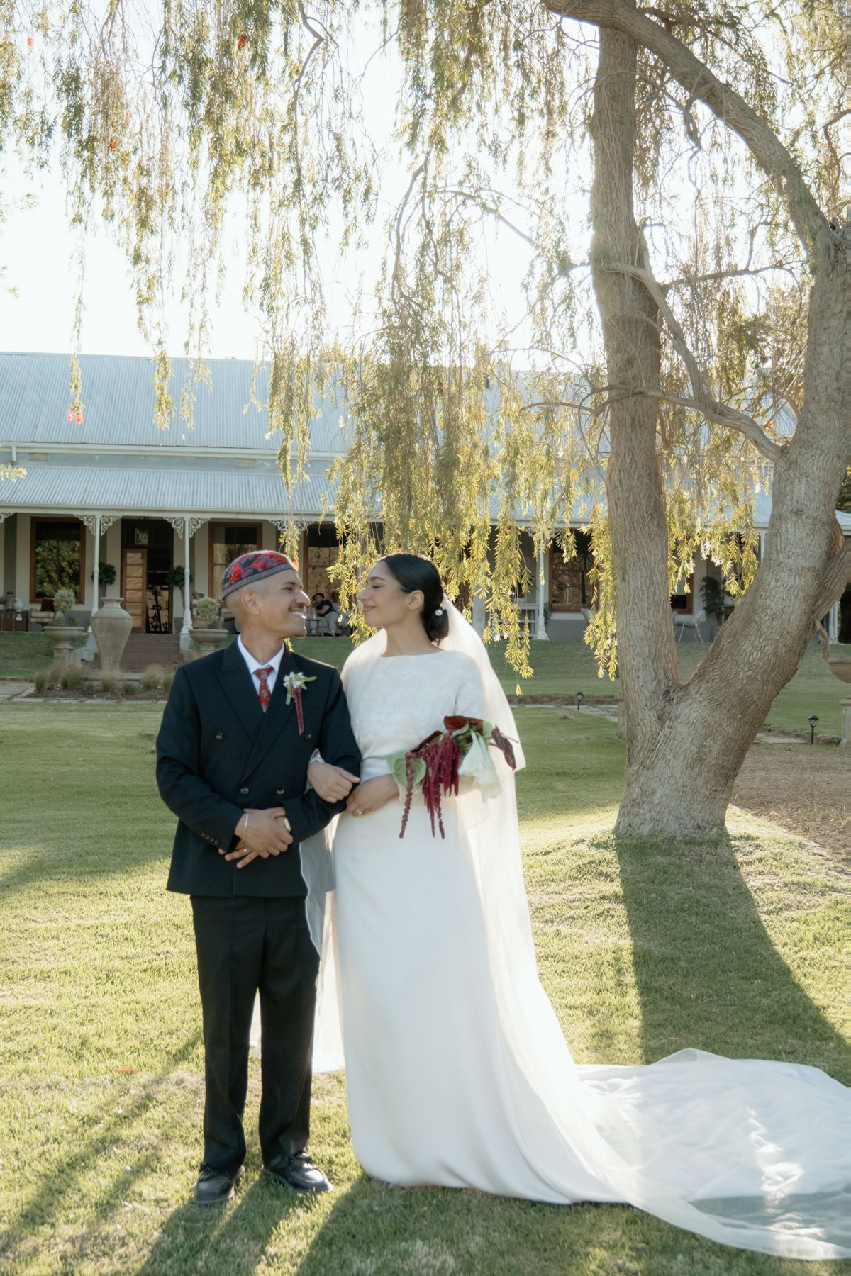 Muslim bride and groom share a smile beneath weeping willow tree at outdoor wedding venue.