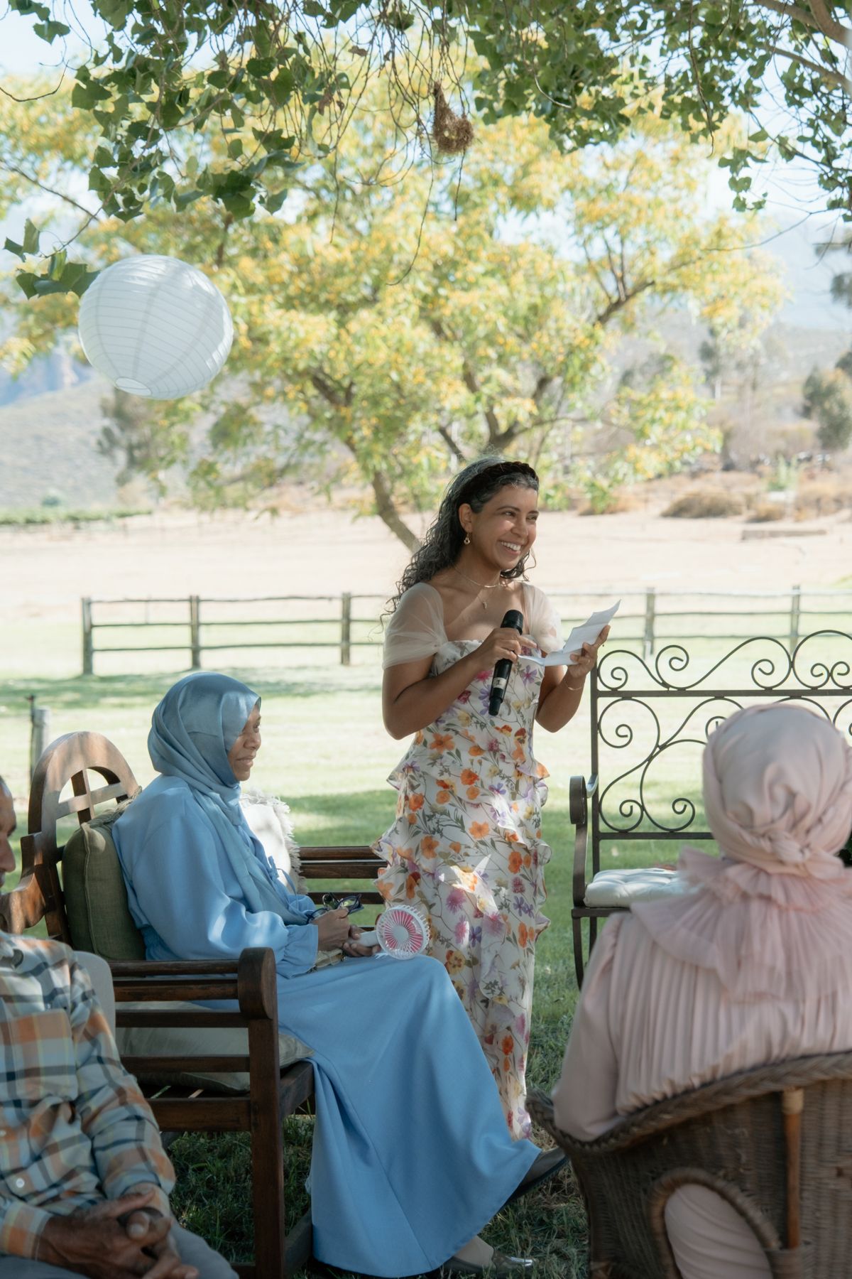 Woman in floral dress gives speech at outdoor wedding while guests in hijabs listen attentively.