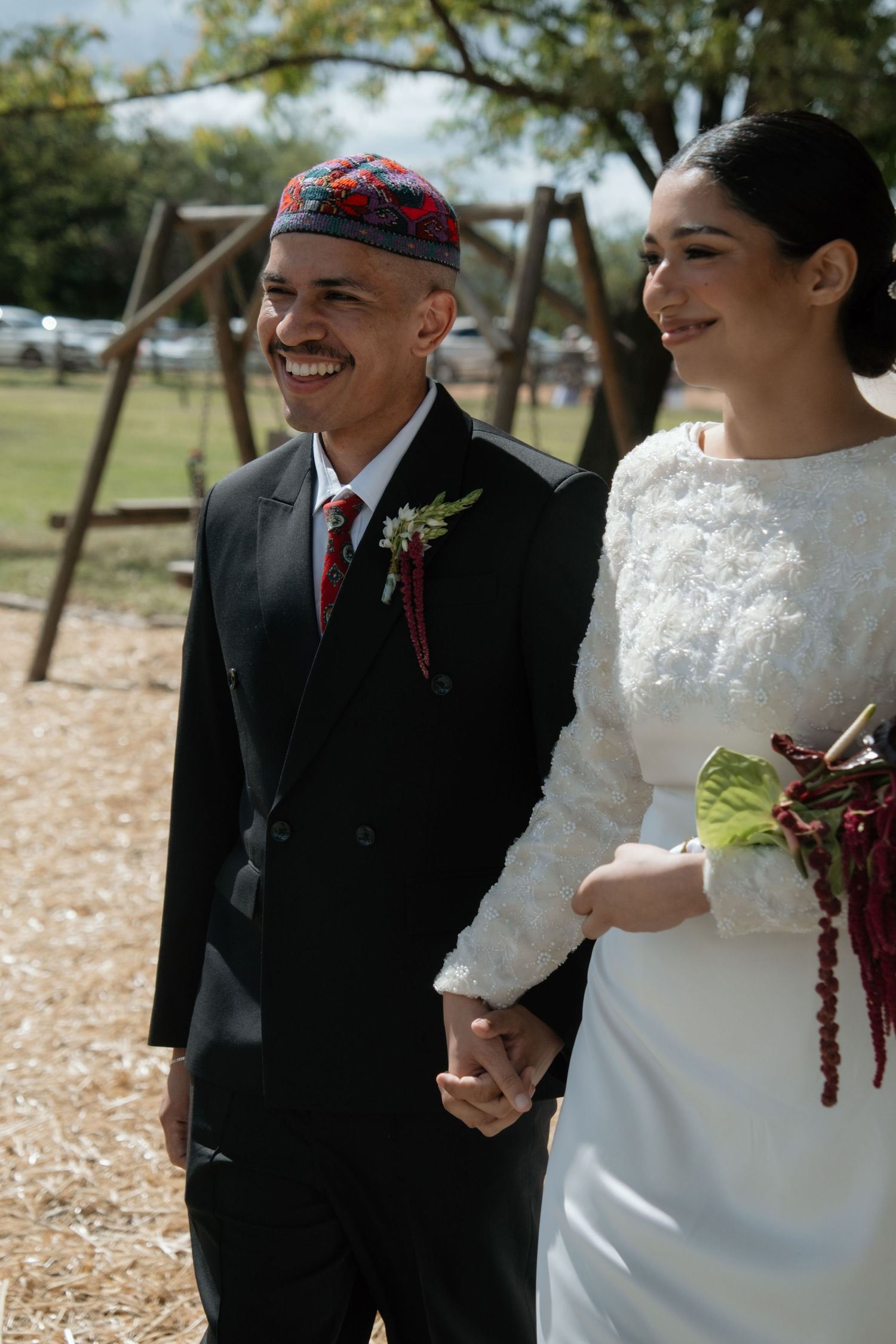Smiling groom in black suit with red kufi cap holds hands with bride in white lace dress outdoors.