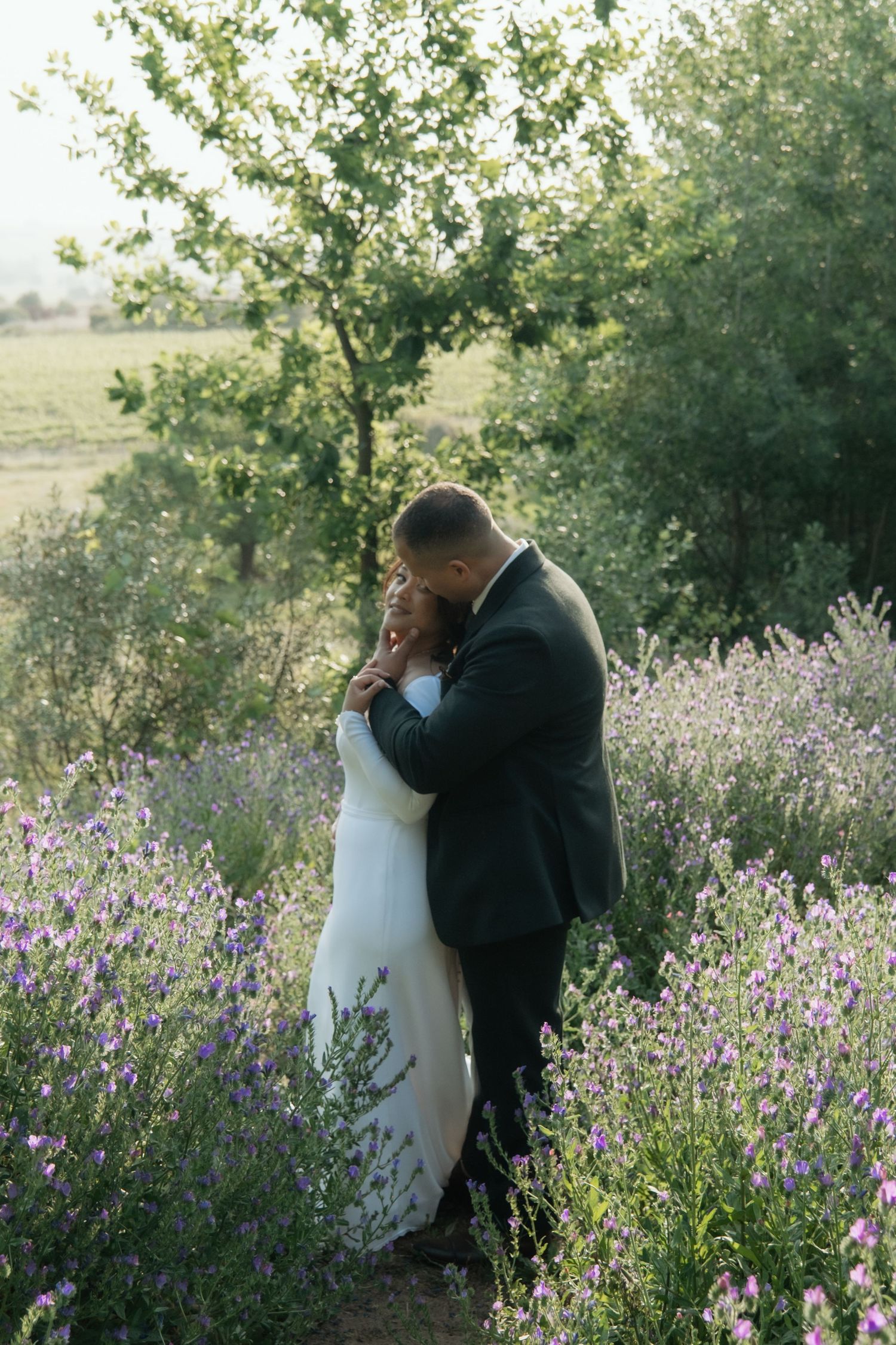 Bride and groom embrace romantically in a lavender field with lush green trees in the background on their wedding day.
