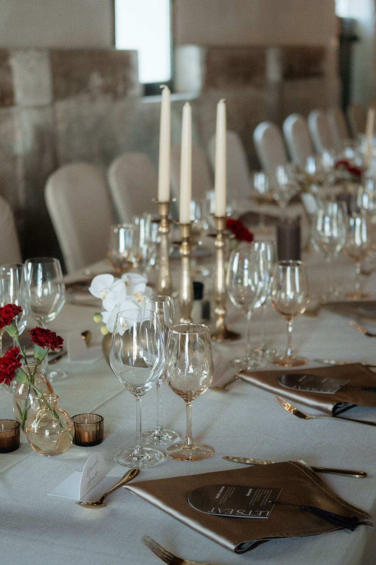 Elegant wedding reception table with crystal glasses, gold candlesticks, and red floral centerpieces.