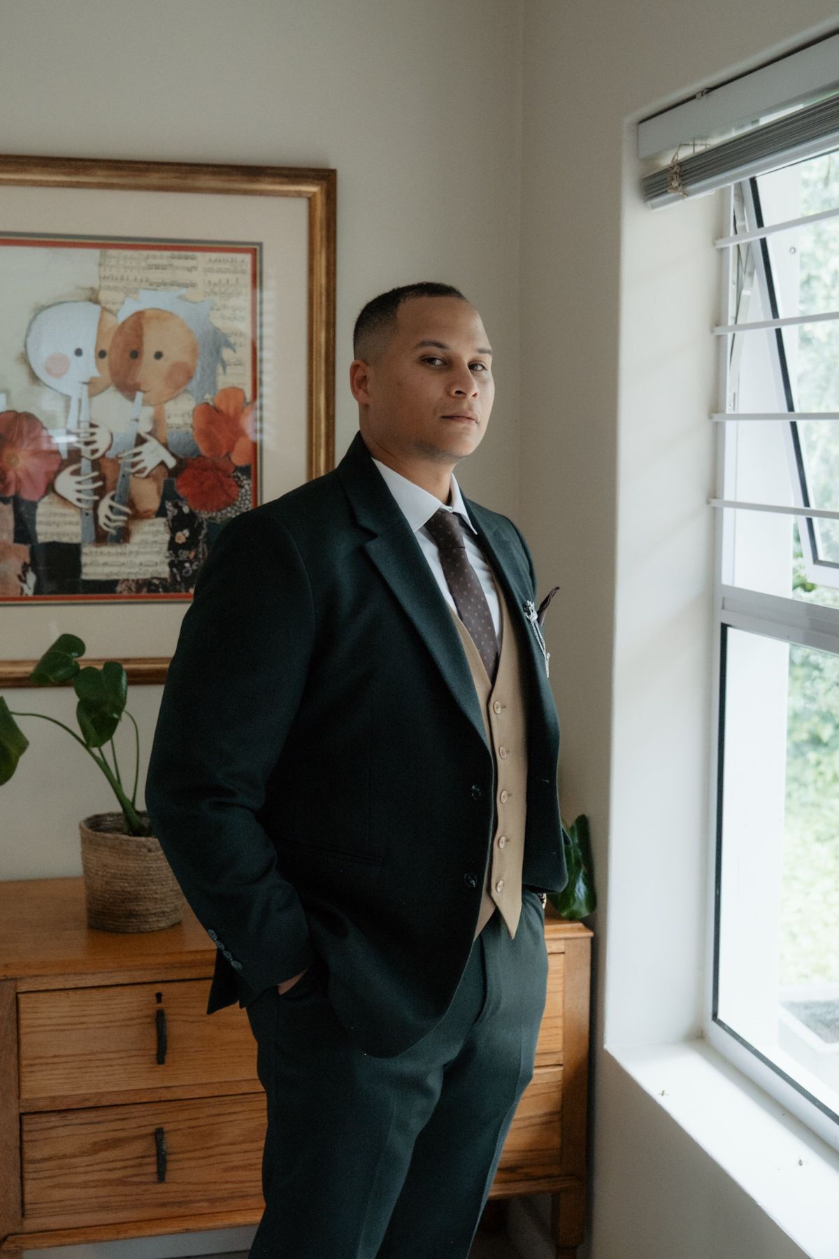 Groom in dark green suit and tan vest standing thoughtfully by a window before his wedding.