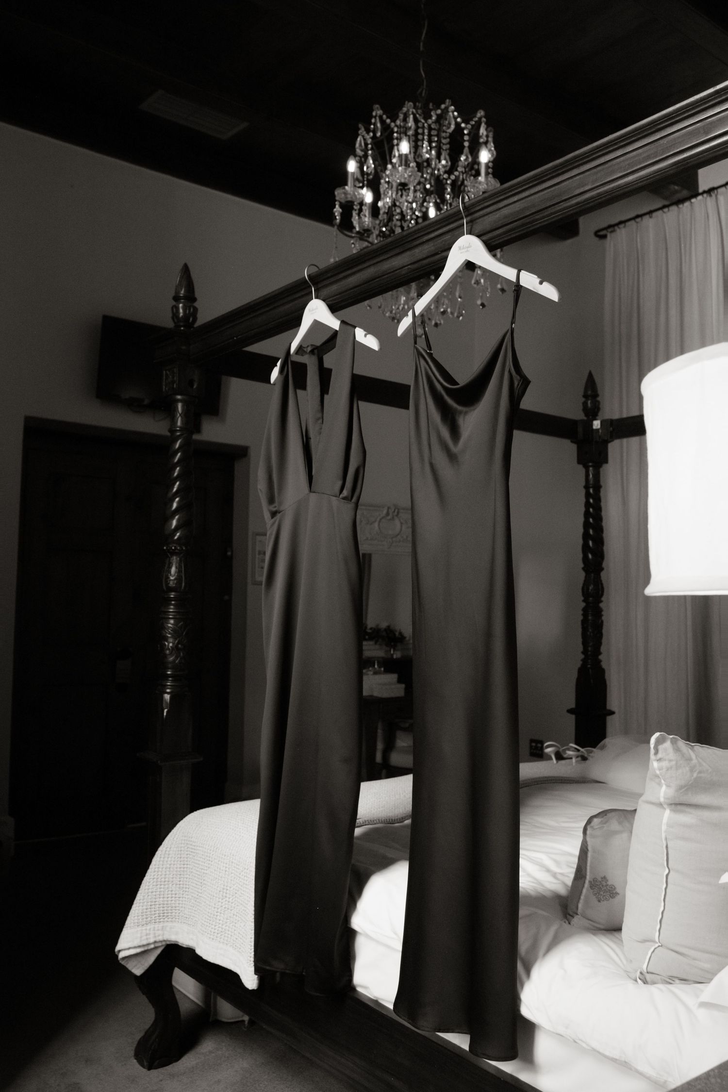 Two elegant dark bridesmaid dresses hanging on a four-poster bed in a black and white photo.