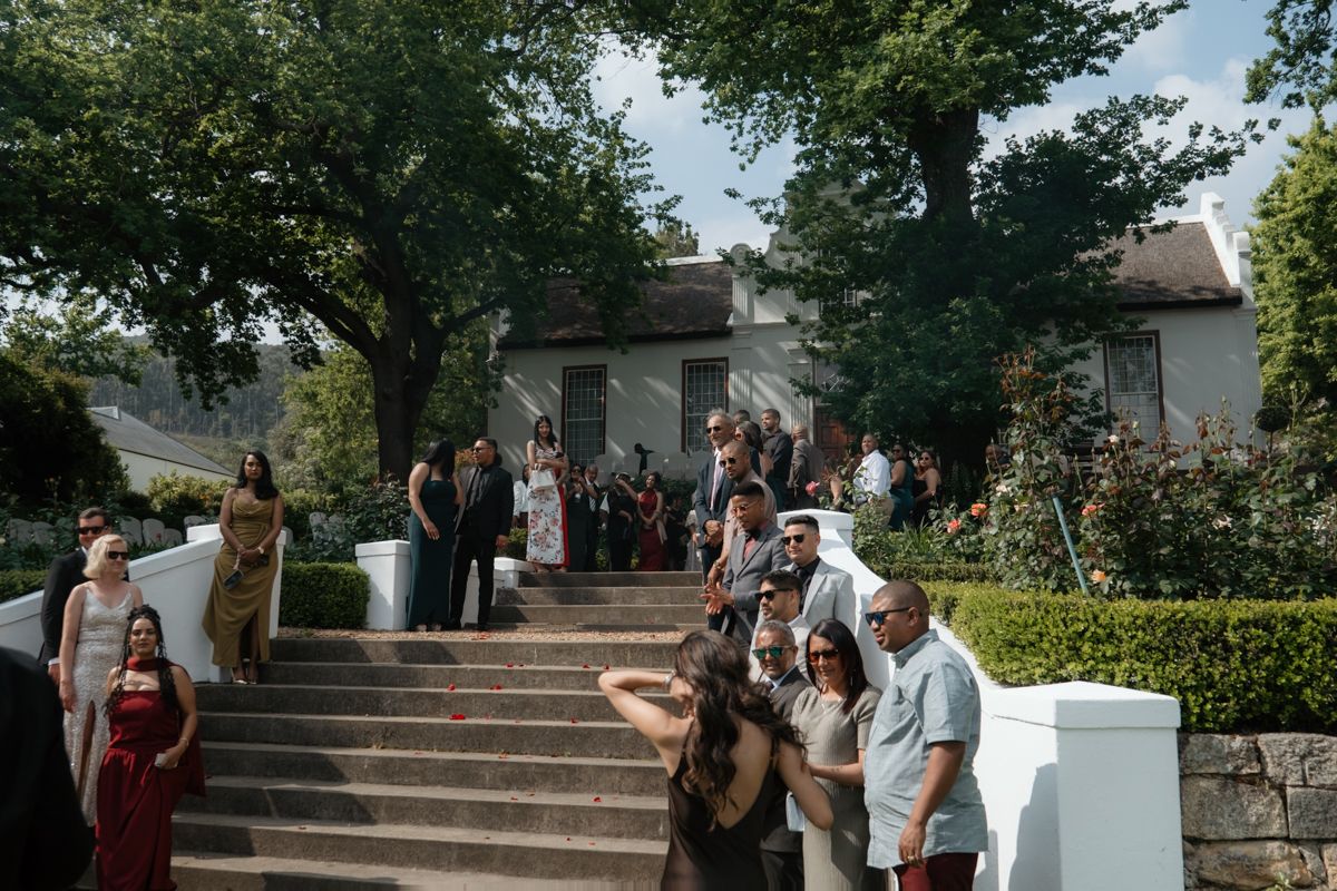 Wedding guests gather and mingle on stone steps outside a charming venue surrounded by trees.