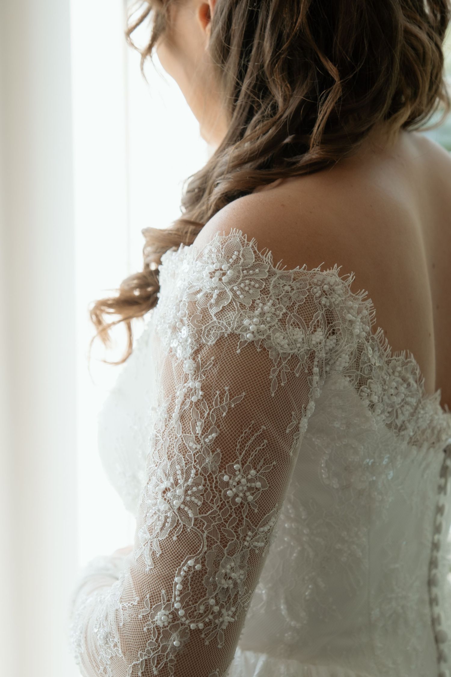 Close-up of bride in elegant off-shoulder lace wedding dress with floral embroidery by a window.