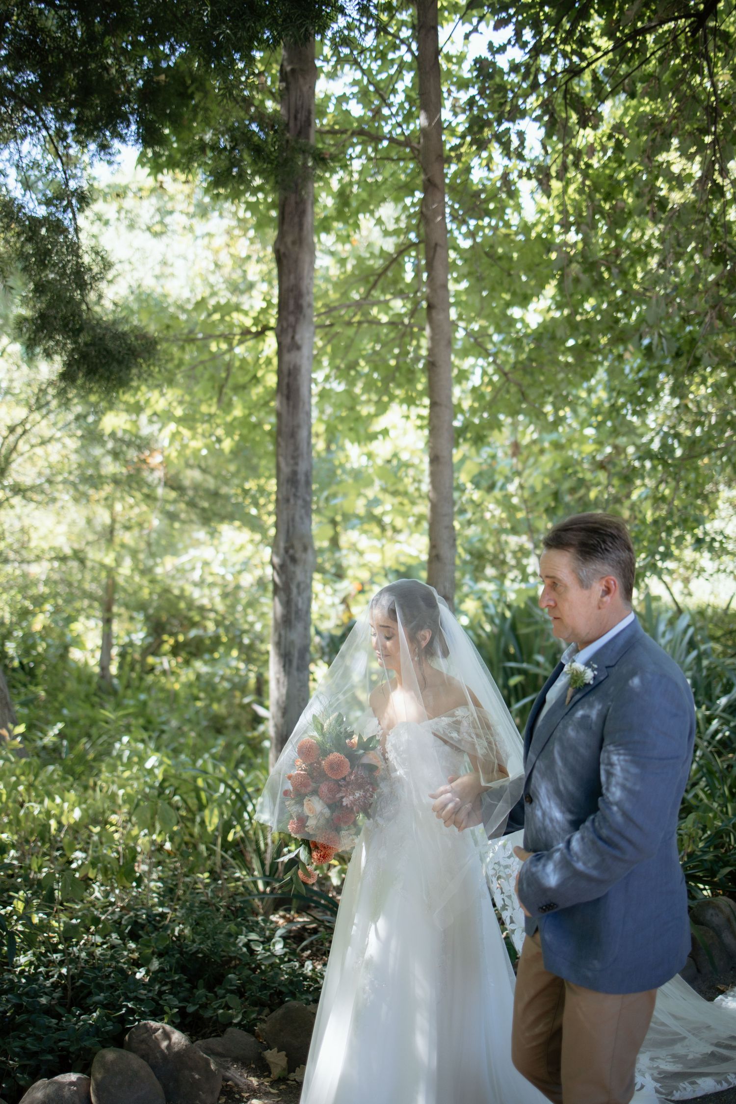 Bride in lace wedding gown with veil holds bouquet beside groom in garden ceremony setting.