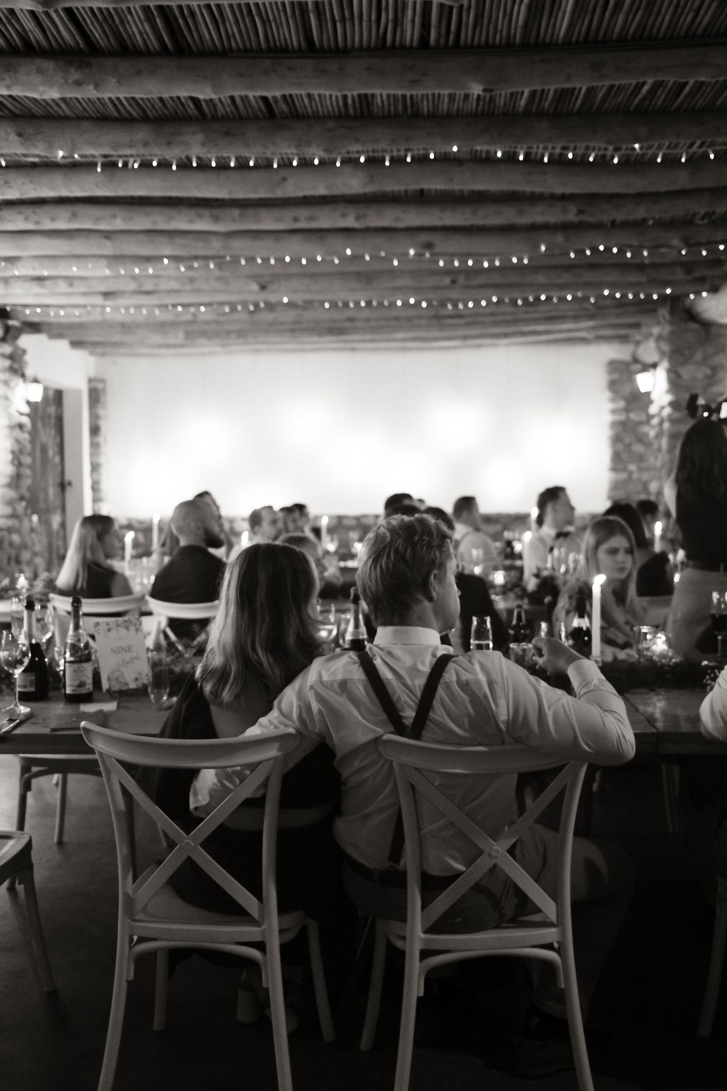 Black and white wedding reception photo of guests seated under rustic thatched ceiling with string lights.