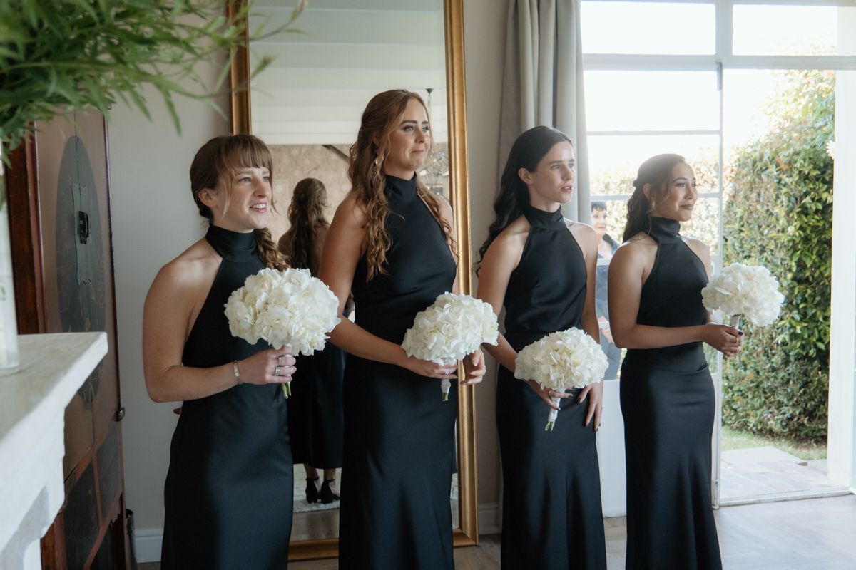 Four bridesmaids in black halter gowns hold white hydrangea bouquets while standing indoors before the ceremony.