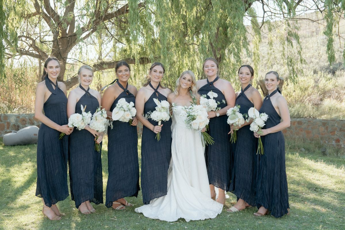Bride with bridesmaids in dark dresses holding white bouquets posing outdoors under a willow tree.
