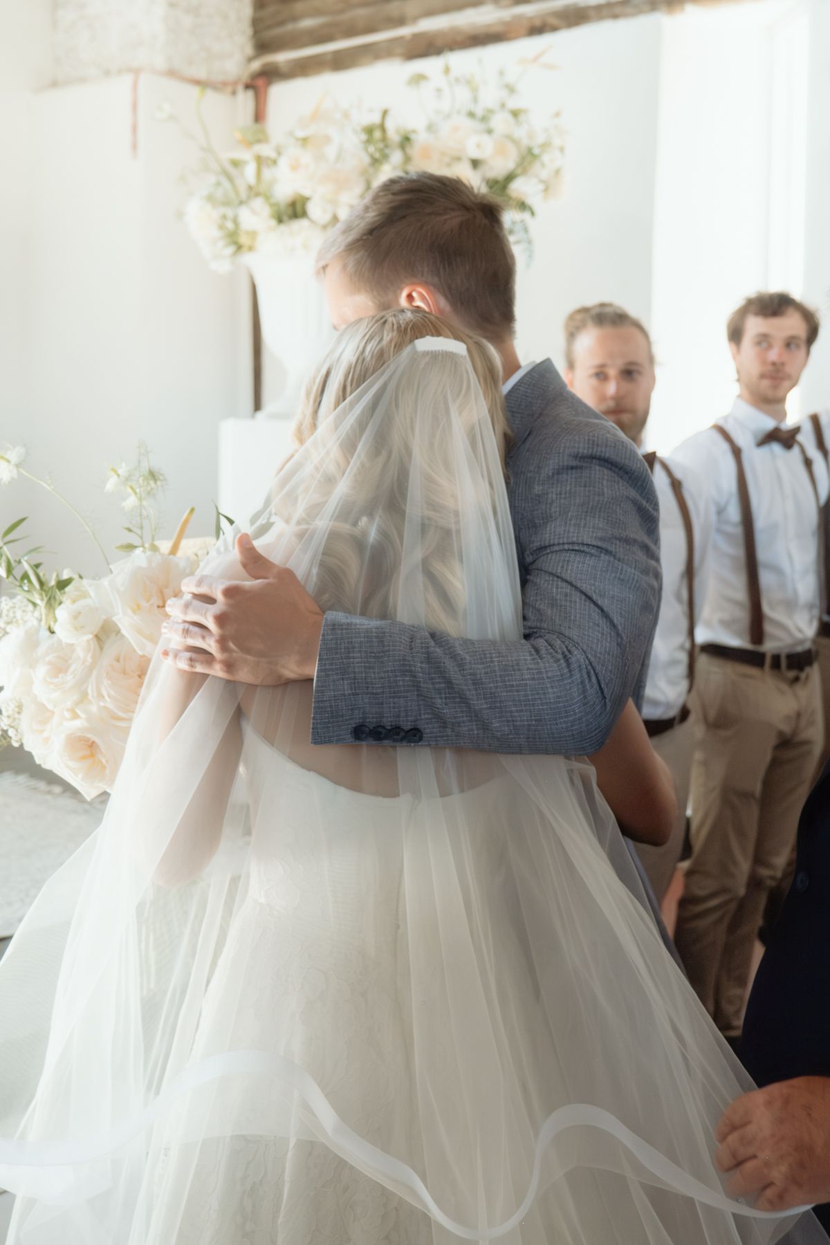 Bride and groom share a tender embrace during wedding ceremony while groomsmen watch in elegant indoor venue.