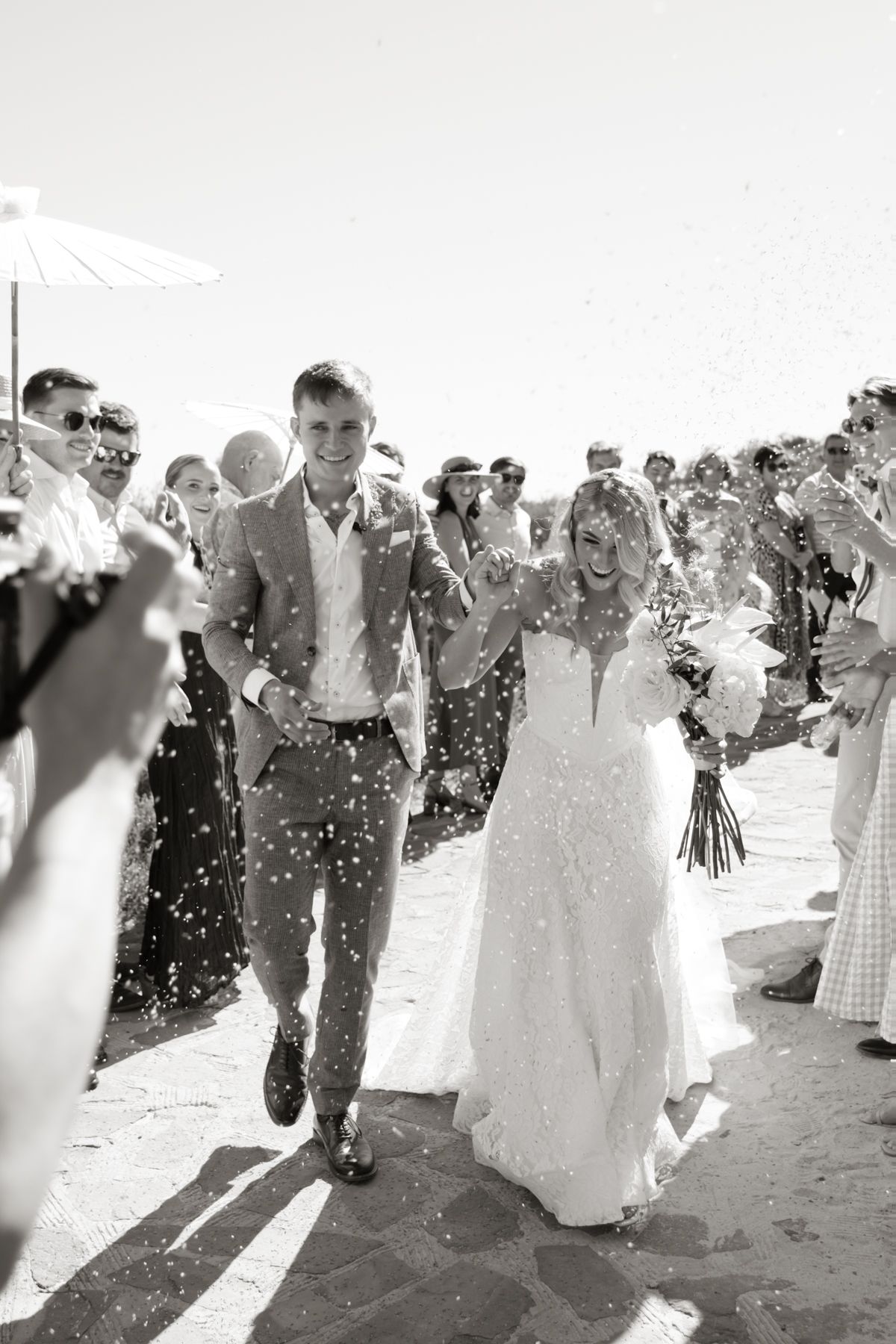 Bride and groom walk through confetti shower surrounded by cheering wedding guests outdoors.