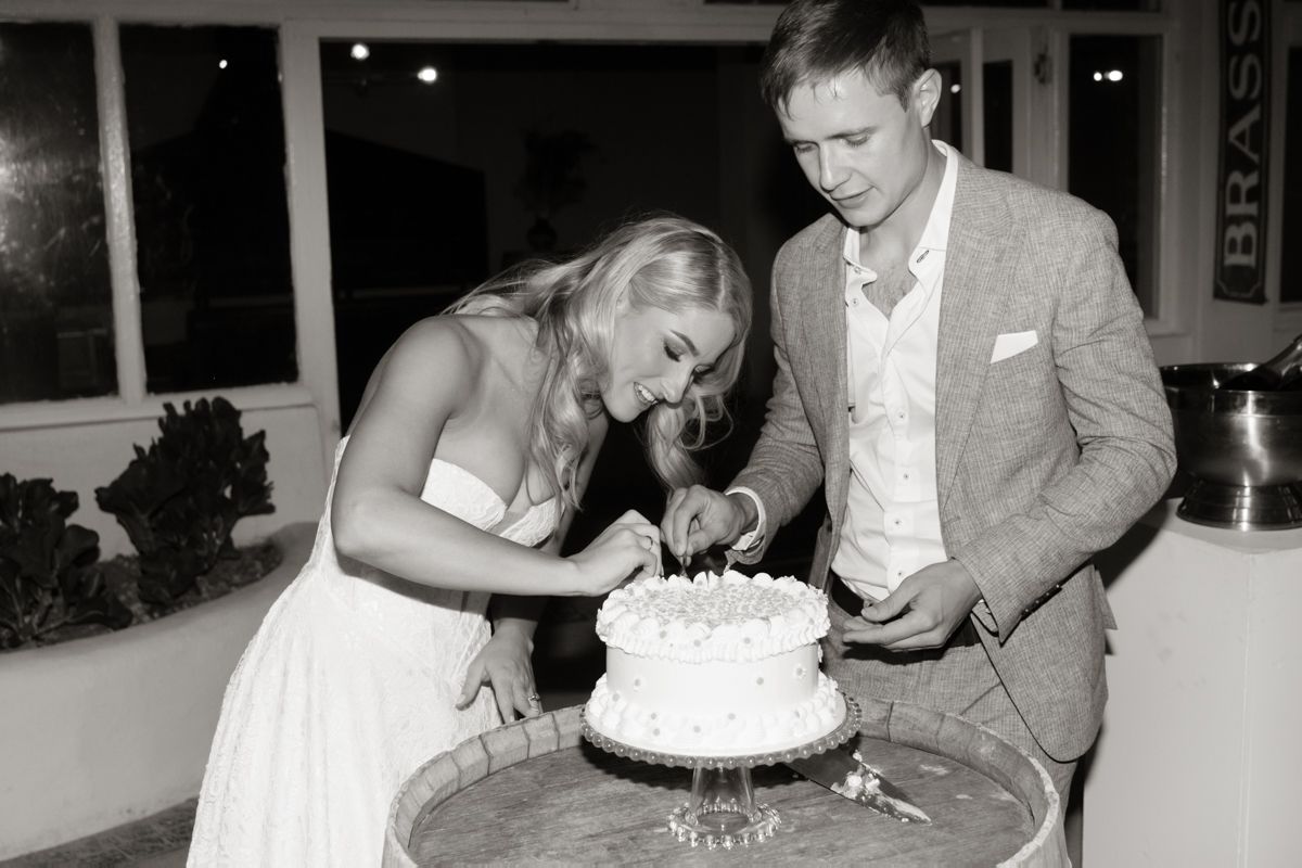 Bride and groom cut their white wedding cake together in a classic black and white reception photo.