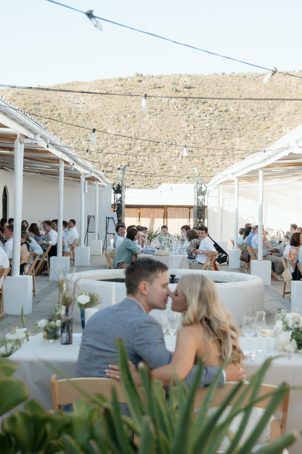 Newlyweds share a kiss at their outdoor Mediterranean wedding reception under string lights with mountain backdrop.