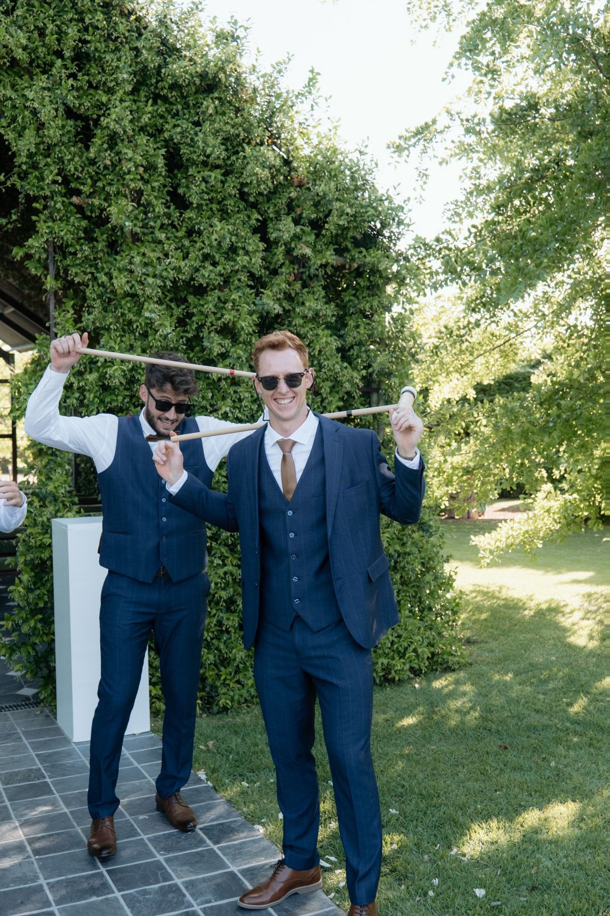 Two groomsmen in navy suits playfully pose with a stick in a lush garden wedding setting.