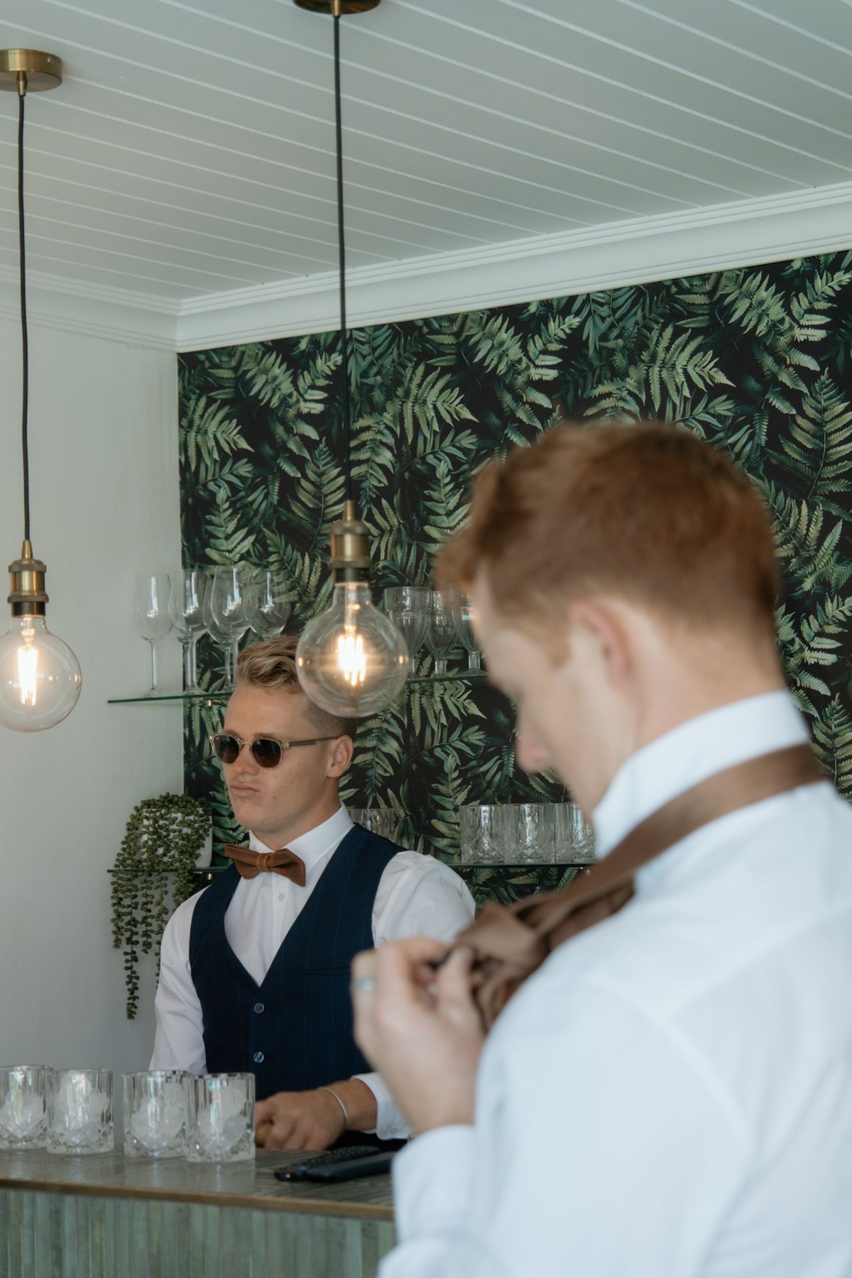 Groomsman in navy vest and bow tie stands at stylish bar with tropical leaf wallpaper.