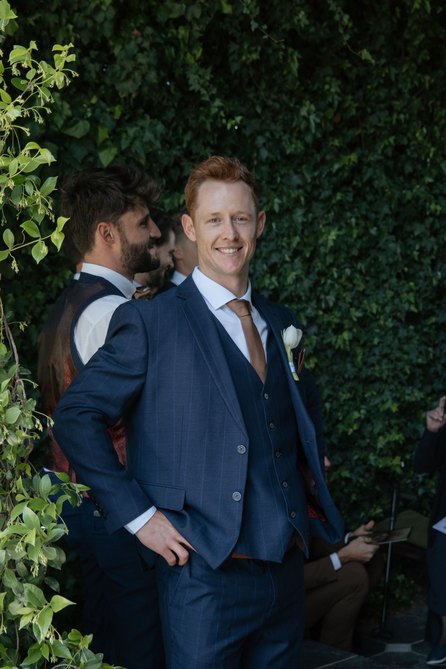 Smiling groom in navy blue three-piece suit with tan tie and white boutonniere stands outdoors against lush green ivy wall.