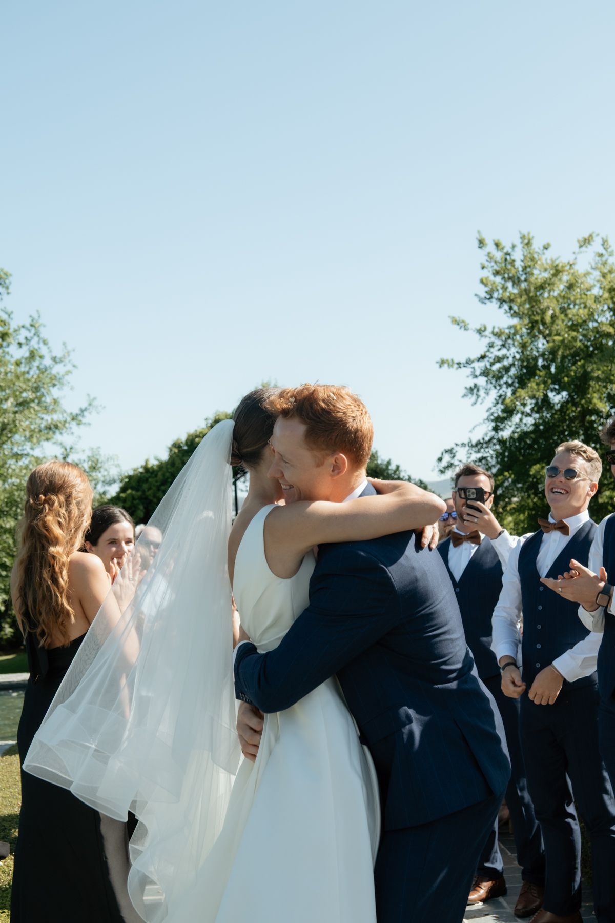 Newlywed couple shares their first kiss while guests cheer at a sunny outdoor wedding ceremony.