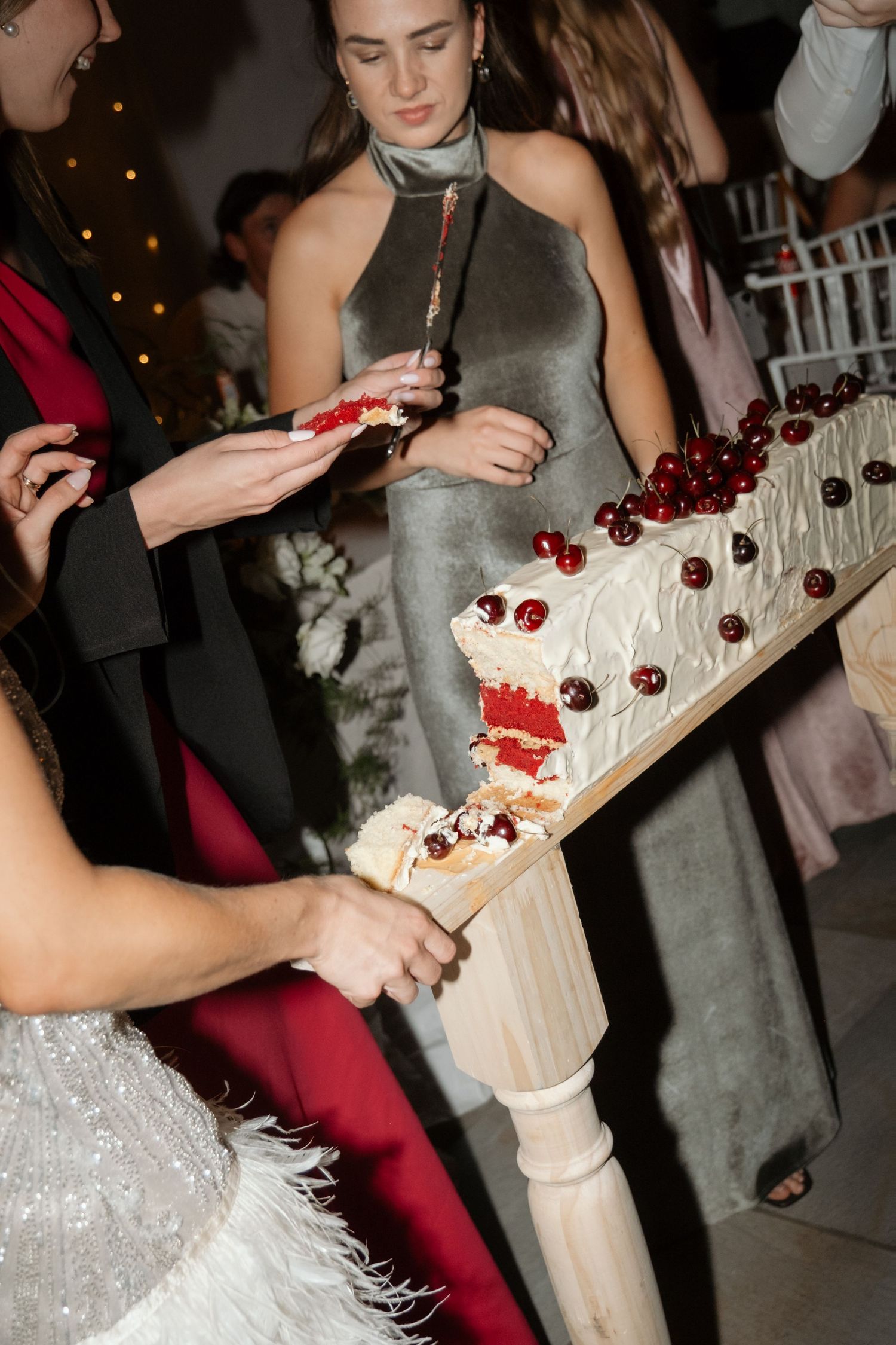 Wedding guests in formal attire serving slices of white cherry-topped cake at elegant reception.