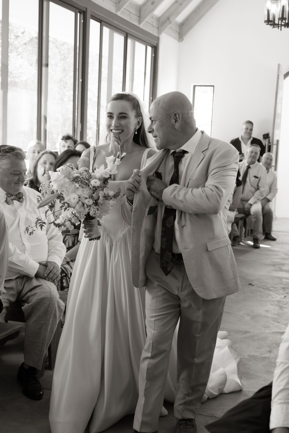 Black and white photo of a bride holding bouquet walking down the aisle with her father at a wedding ceremony.