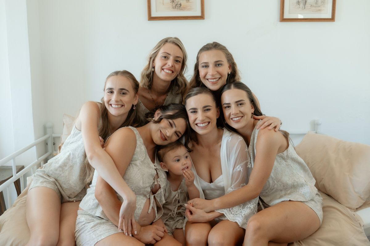 Seven young women smiling and posing together on a couch in white outfits, sharing a joyful group moment.