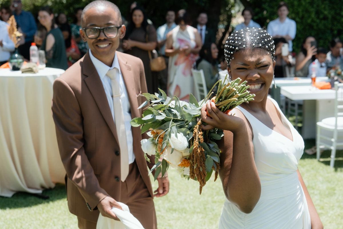 Joyful newlywed couple walking after outdoor wedding ceremony, groom in brown suit, bride in white dress holding bouquet.