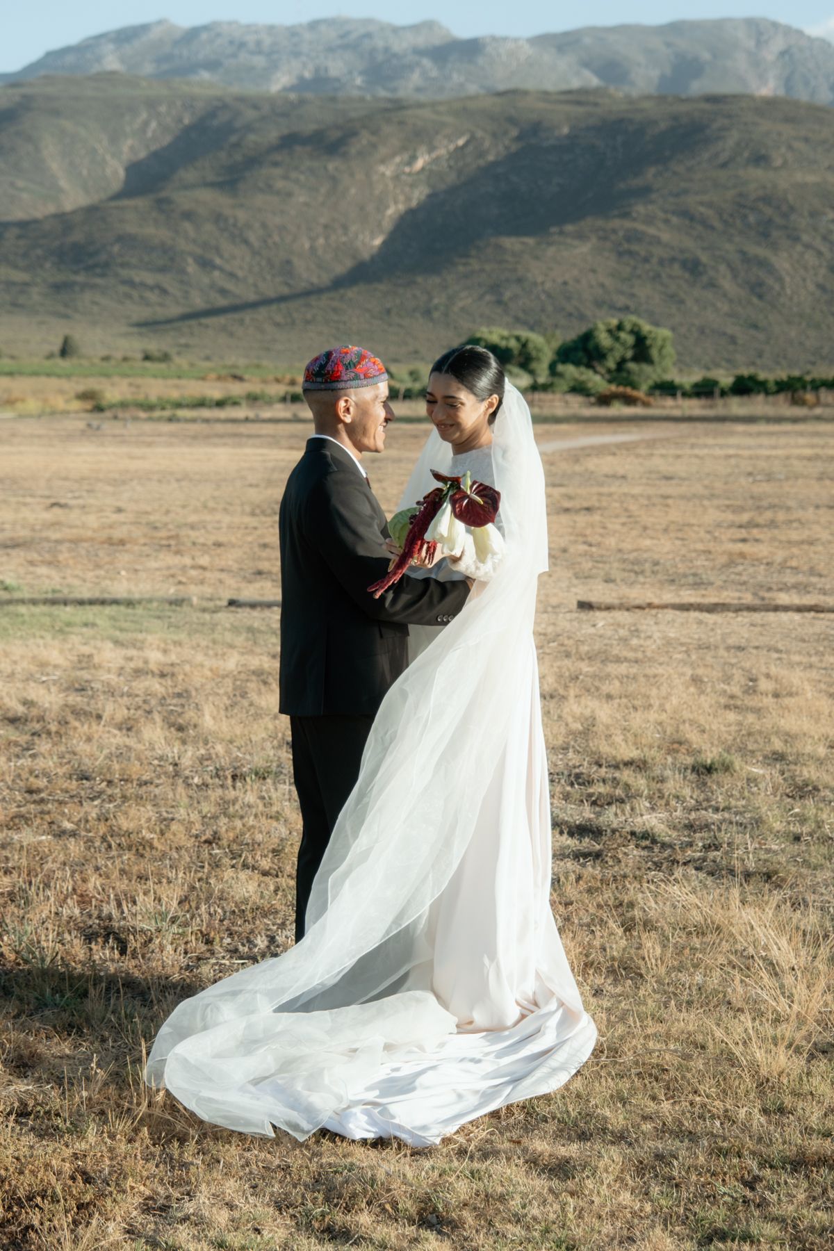 Bride and groom standing face to face in a golden field with dramatic mountain scenery at sunset.