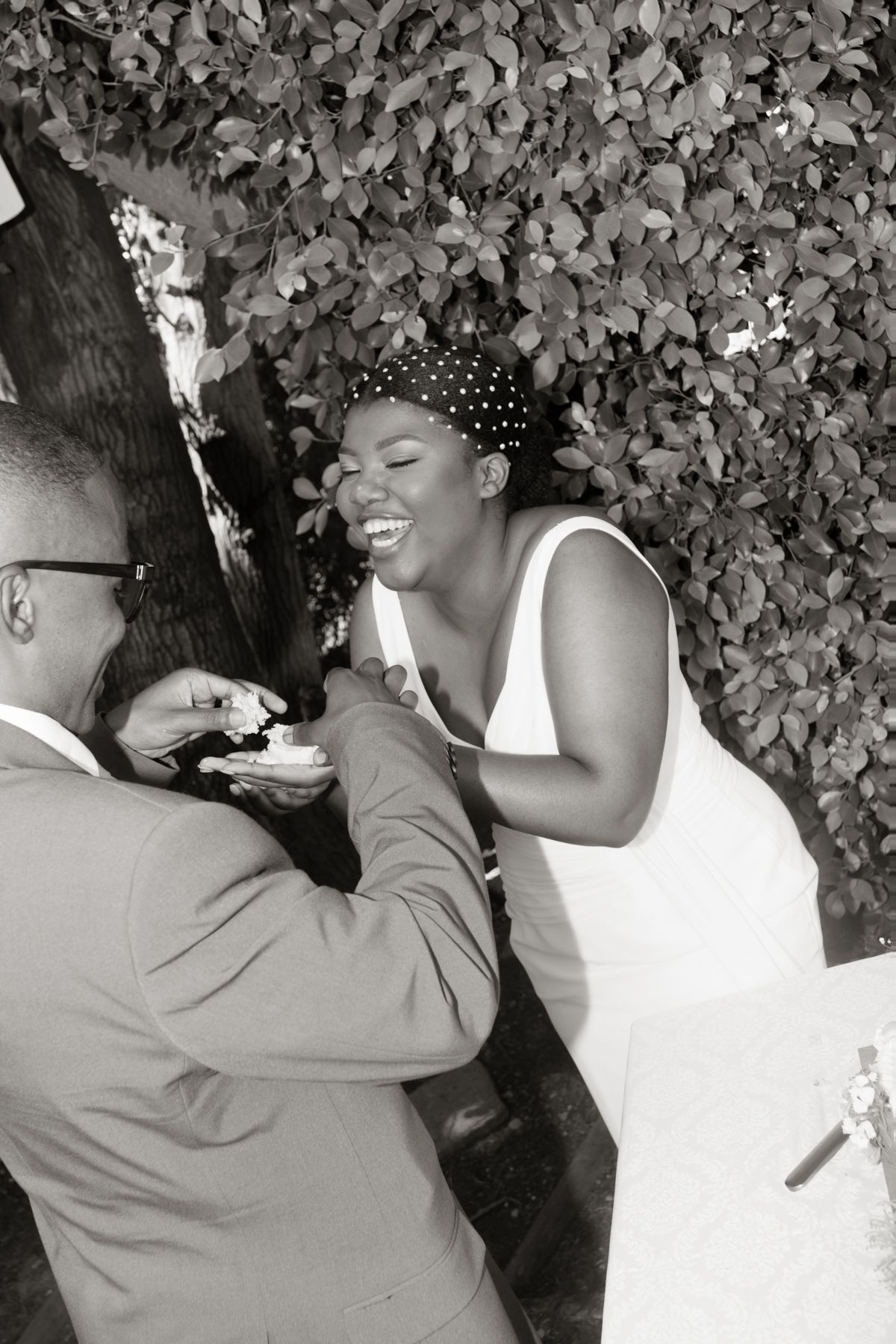 Bride in white dress laughing joyfully as groom feeds her wedding cake outdoors in black and white photo.
