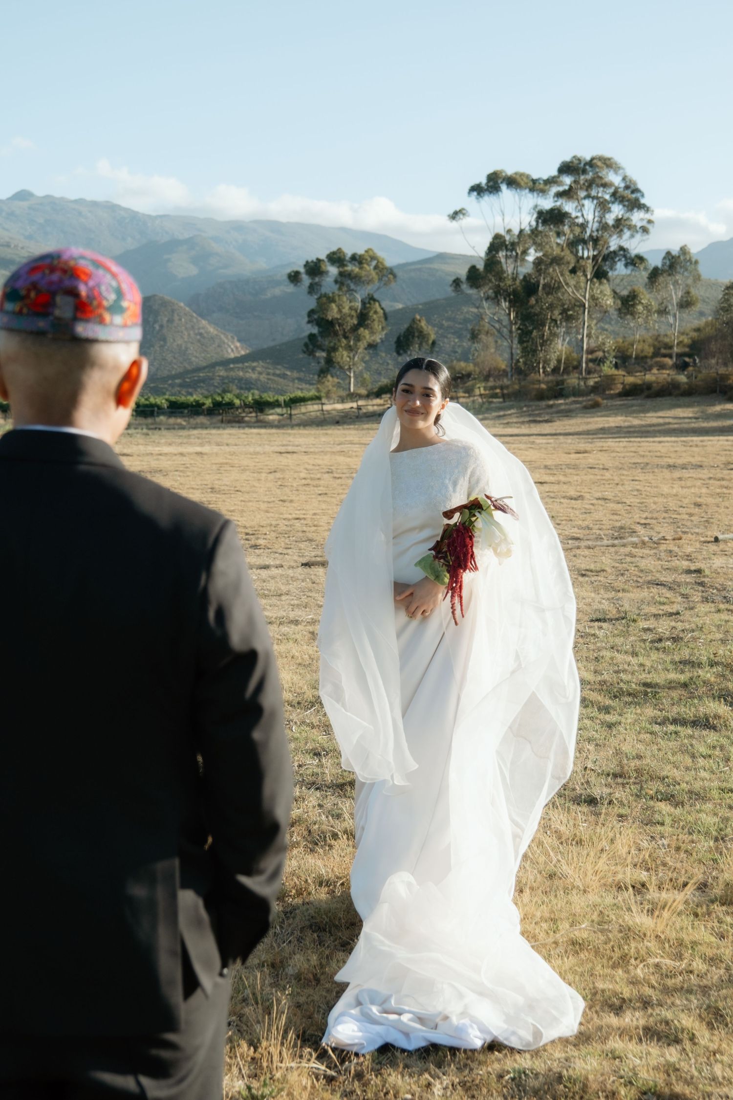 Bride in flowing white veil holds red bouquet facing groom in black suit at outdoor mountain wedding.