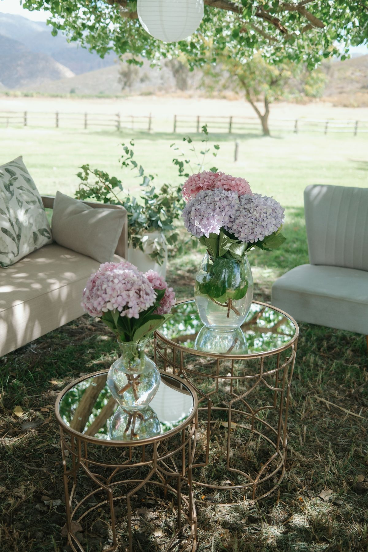 Pink hydrangea floral arrangements in glass vases on gold mirrored tables at outdoor wedding.