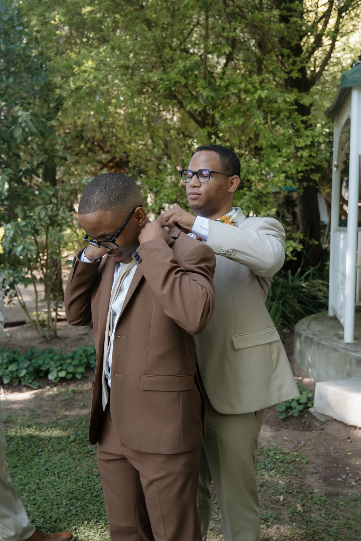 A groomsman helps adjust the jacket of a man in a brown suit at an outdoor wedding venue.