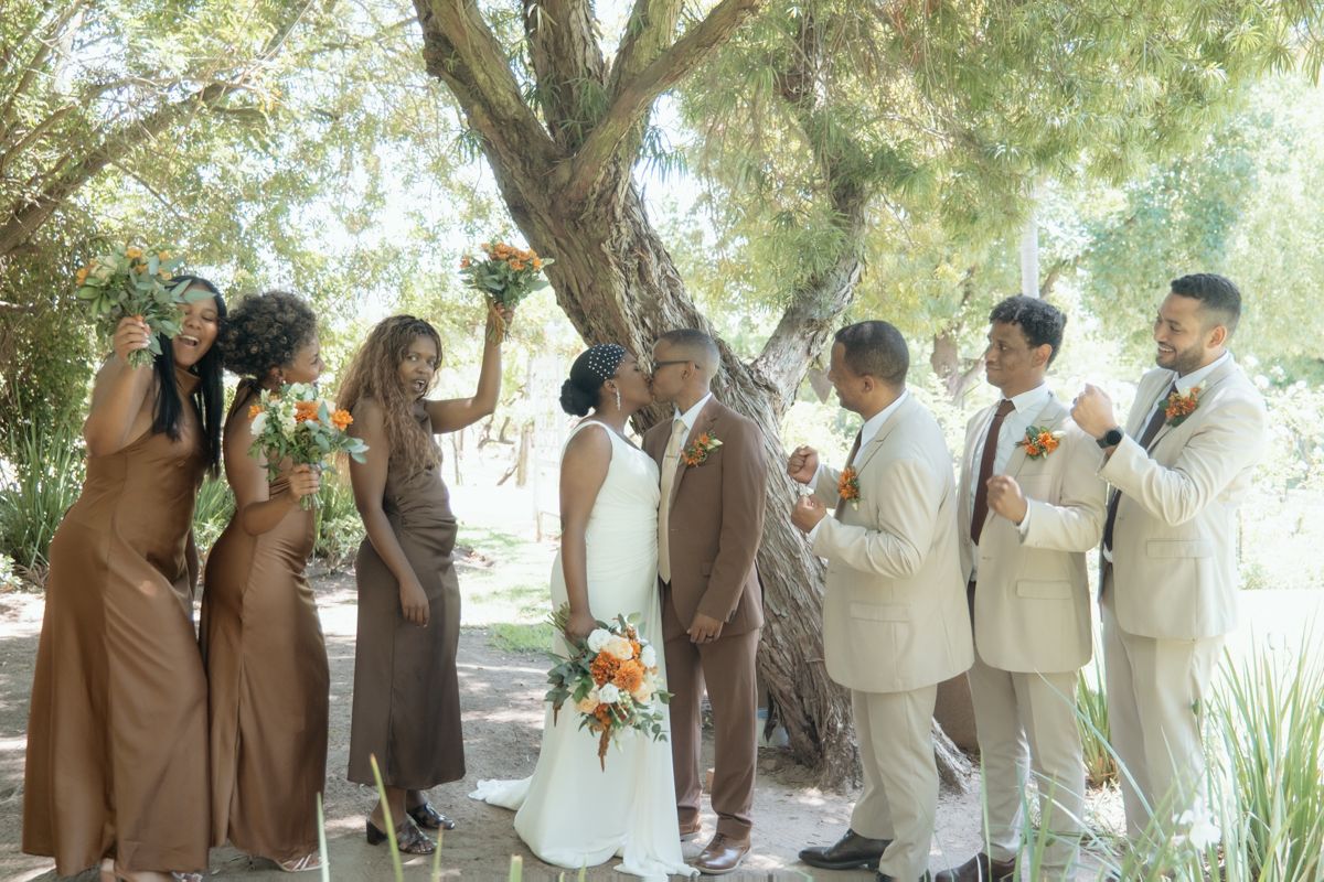 Wedding party celebrates under a tree, bridesmaids in brown gowns and groomsmen in tan suits cheering for kissing couple.