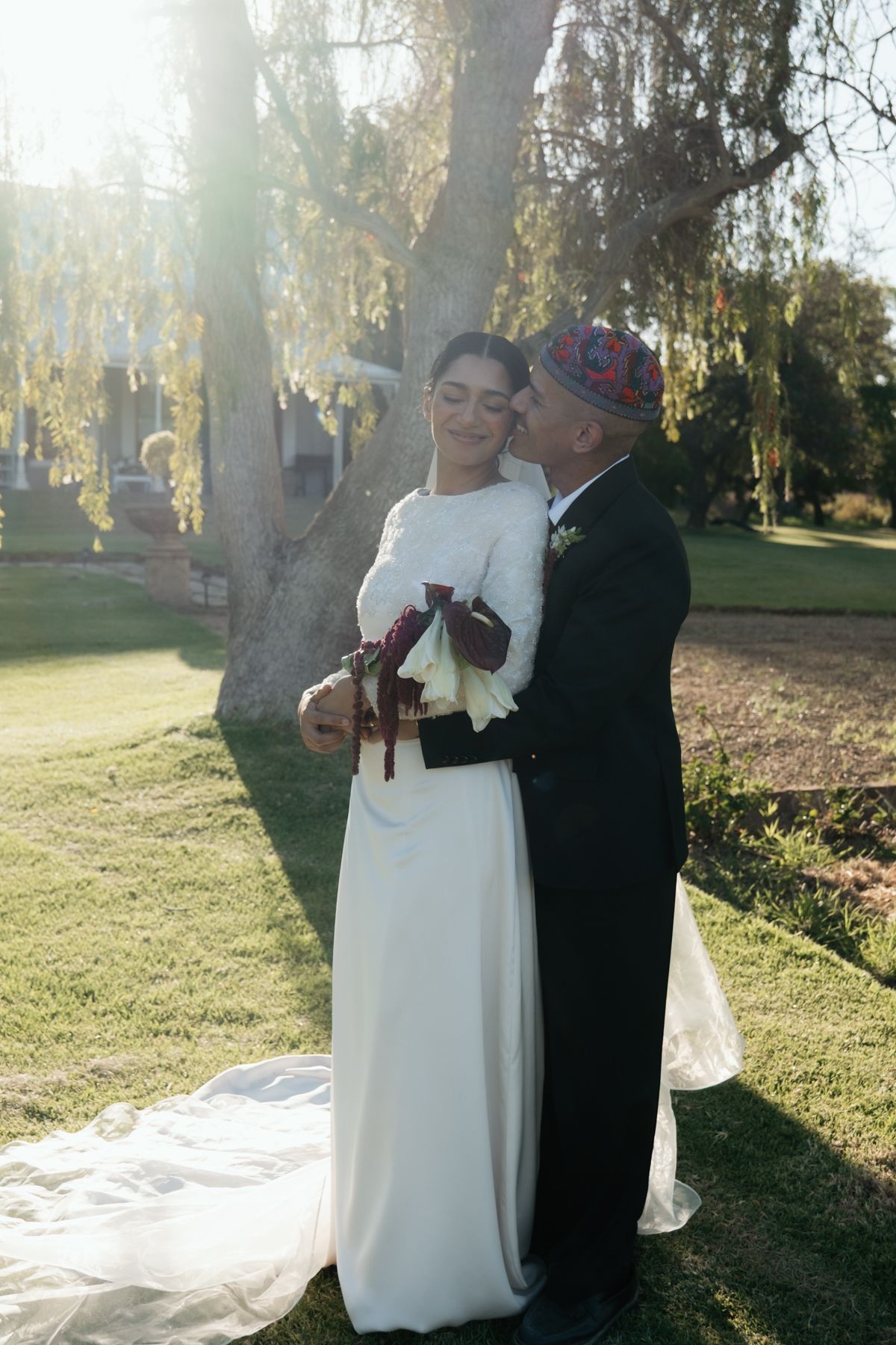 Groom in embroidered skullcap embraces bride in white gown holding red and white bouquet under sunlit tree.