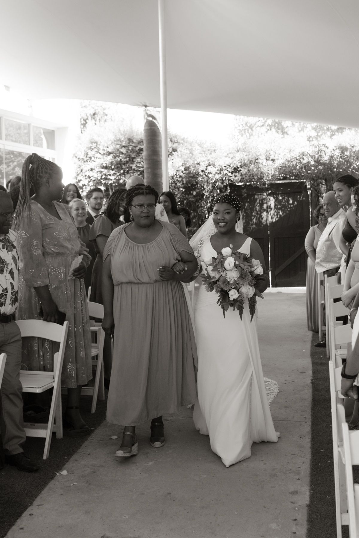 Black and white photo of a bride walking down the aisle with her mother at an outdoor wedding ceremony.