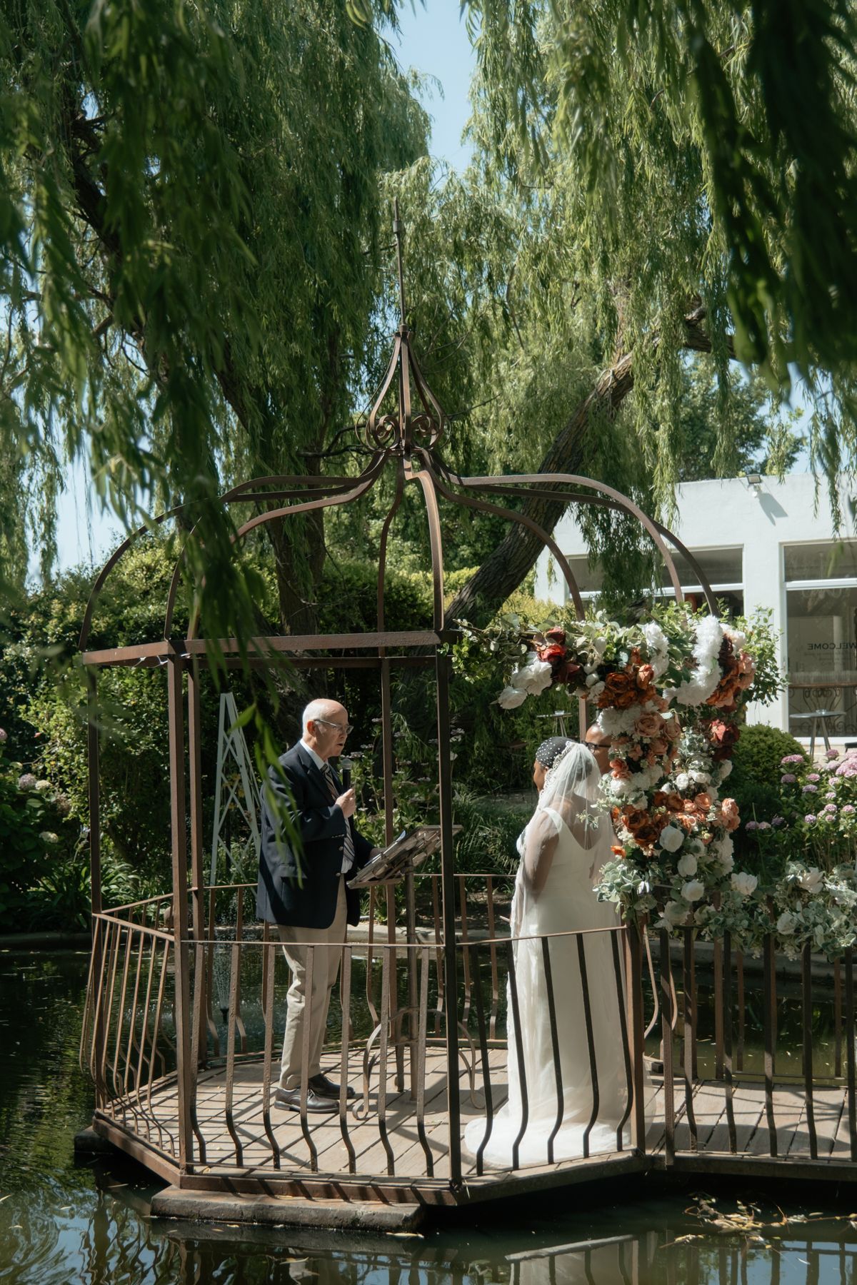 Bride and officiant stand in ornate iron gazebo over water surrounded by lush weeping willow trees.