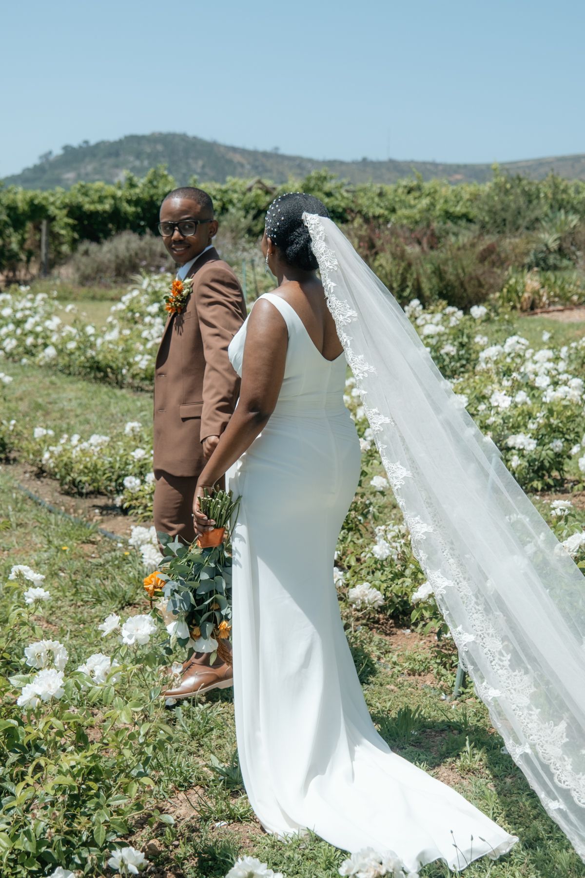 Newlyweds holding hands in a blooming white rose garden with a scenic mountain backdrop on their wedding day.