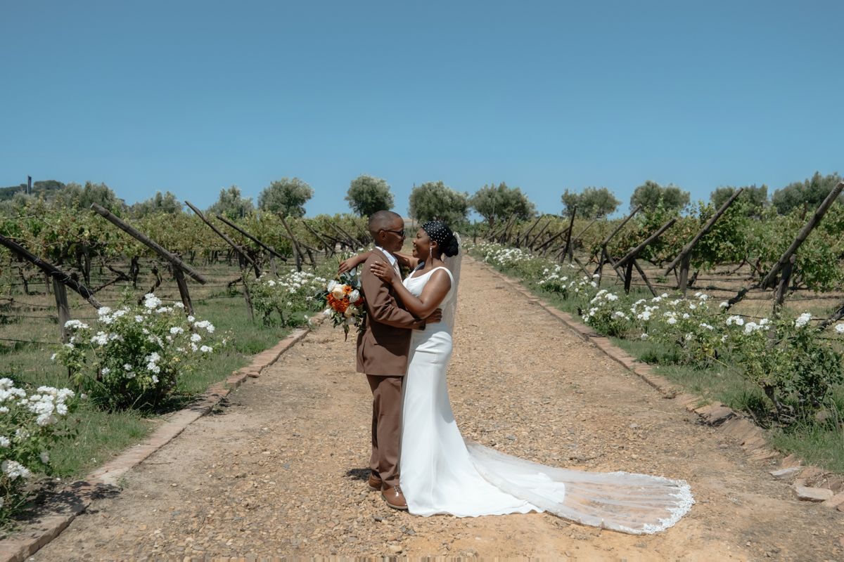 Bride and groom embrace in a vineyard aisle lined with white roses under a clear blue sky.