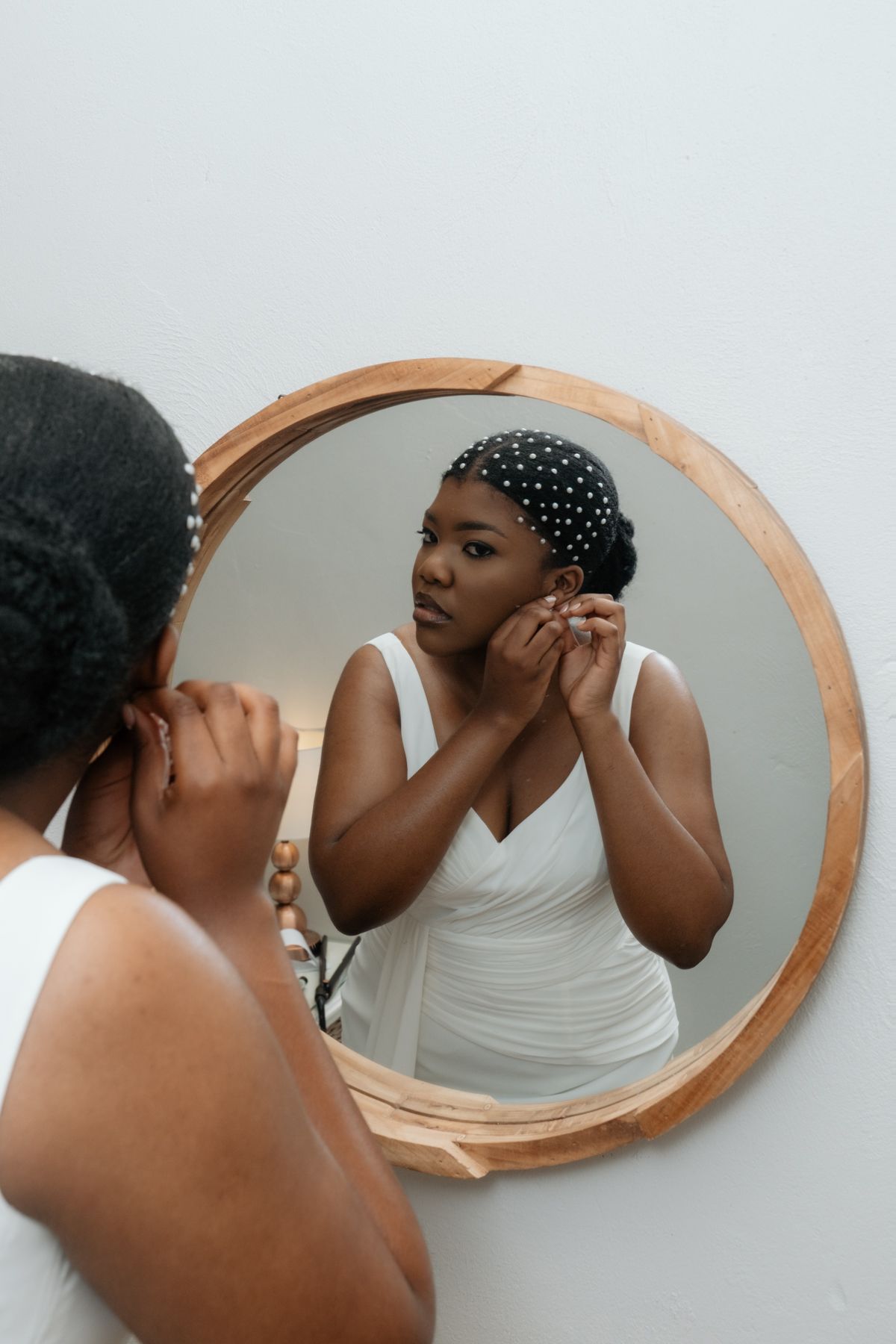 Woman in white dress adjusting earring while looking in round wooden mirror, wearing polka dot headband.