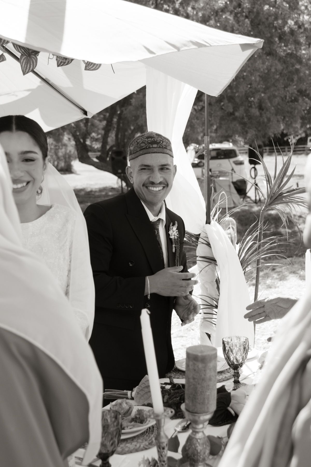 Black and white photo of smiling groom in traditional cap at an outdoor wedding reception under white umbrellas.