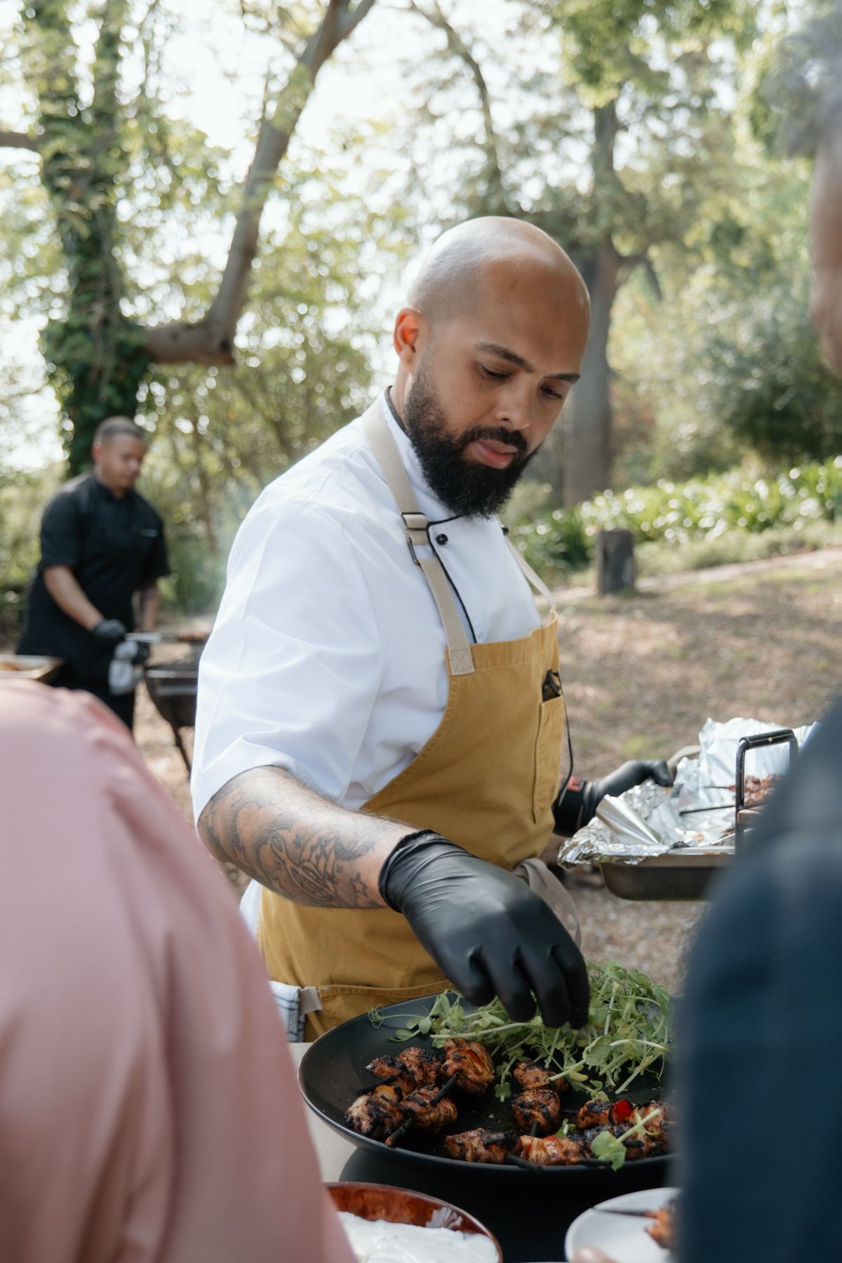 Bald bearded chef in yellow apron plating grilled meat with greens at outdoor wedding.
