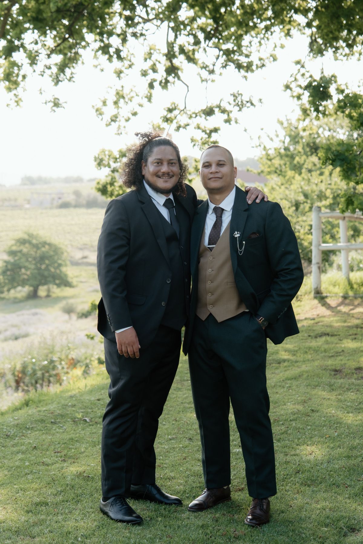 Two groomsmen in dark suits pose together outdoors at a wedding venue with green countryside views.