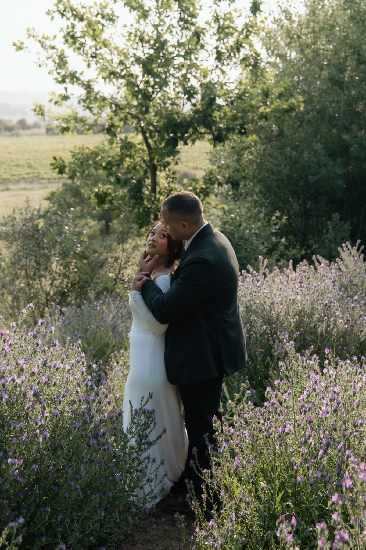 Bride and groom share romantic embrace in lavender field with lush green trees in golden hour light.