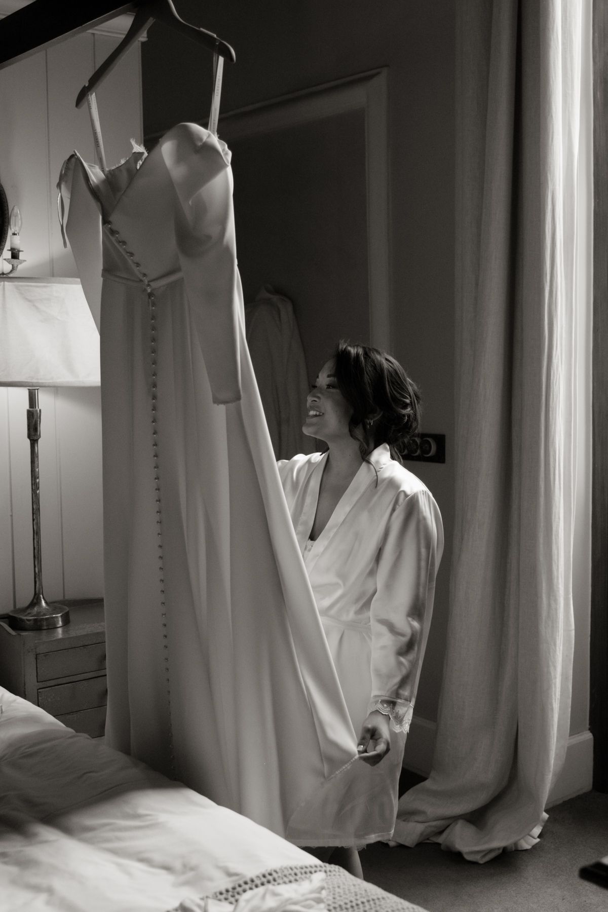 Bride in white robe smiles at her wedding gown hanging in bedroom in black and white photo.