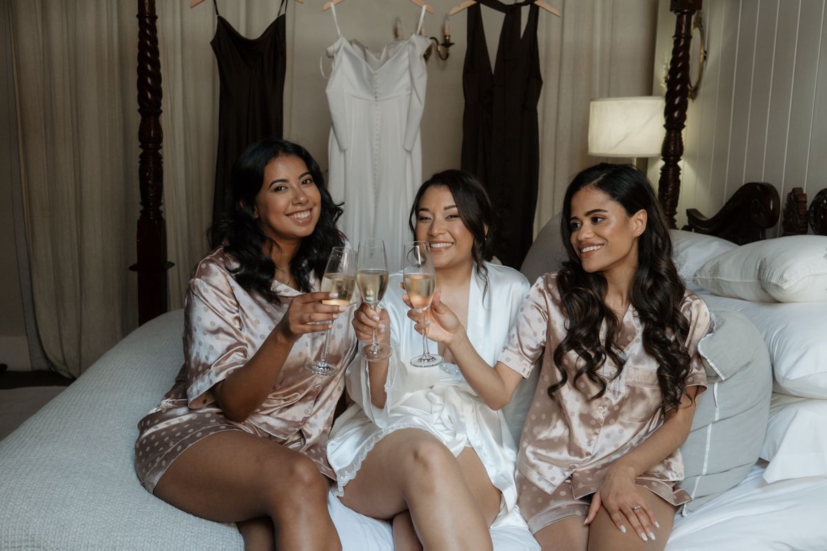 Three bridesmaids in satin robes toast champagne while sitting on bed with wedding dress in background.