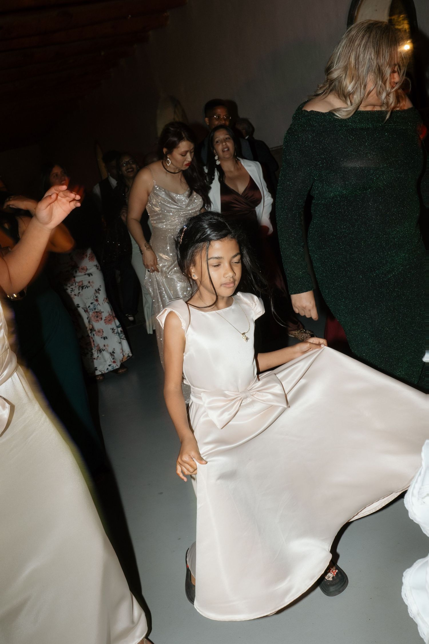 Young flower girl in white satin dress twirls joyfully on the wedding reception dance floor surrounded by guests.