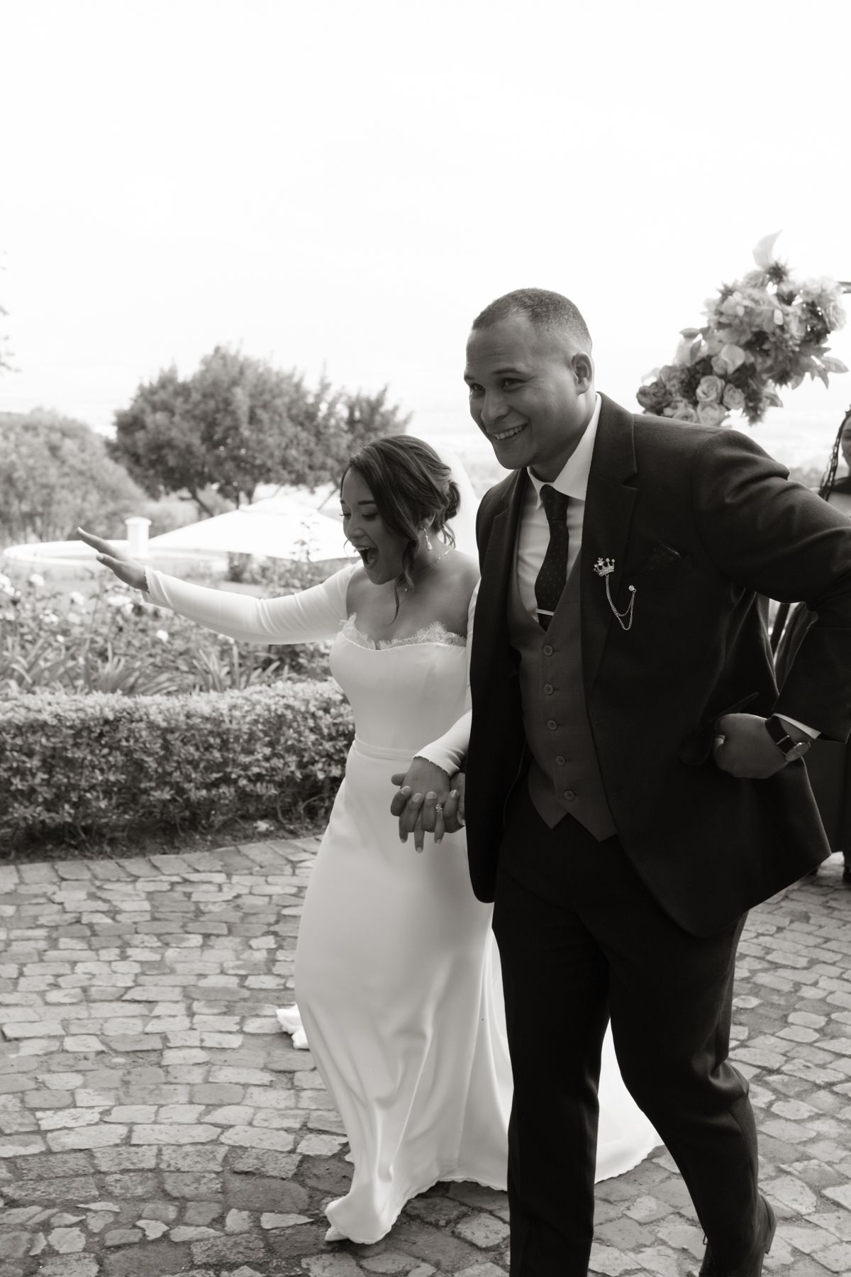 Black and white photo of smiling newlyweds holding hands and walking together after outdoor wedding ceremony.
