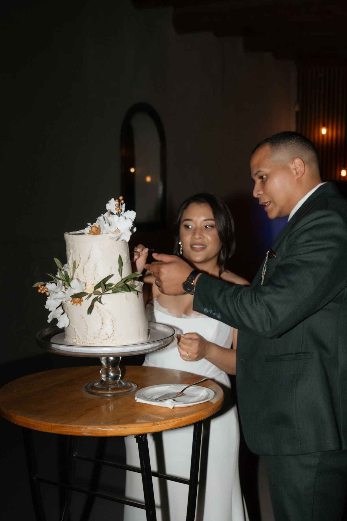 Bride and groom cutting two-tier white wedding cake decorated with white flowers at evening reception.