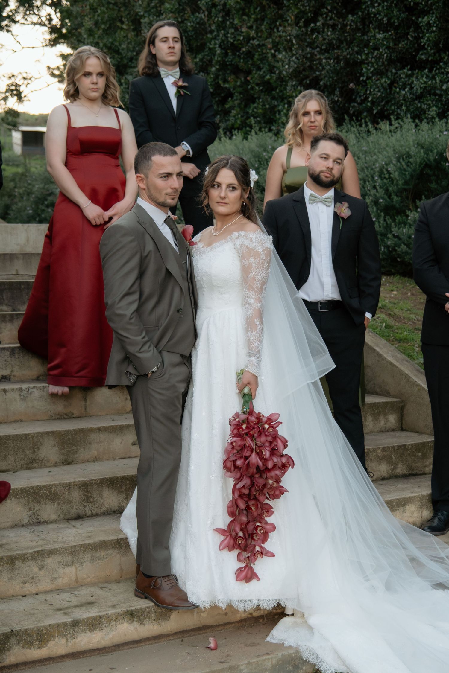 Wedding party portrait on stone steps with bride holding red cascading bouquet and groom in grey suit.